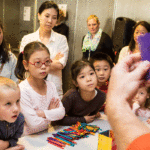 Many youths look on as a person holds up a popsicle stick construction