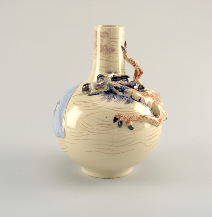 Carved dragon vase (first kiln - in red); cast, cream colored stoneware body with hand-modeled dragon encirling the shoulder and neck in high relief with one leg freestanding. Cobalt blue underglaze and red overglaze decoration; gilt highlights. On reverse, fan-shaped panel of pale underglaze blue painted in overglaze brown with birds flying above cattails and rushes. Allover highly stylized, partially gilded wave pattern. Clear glaze.