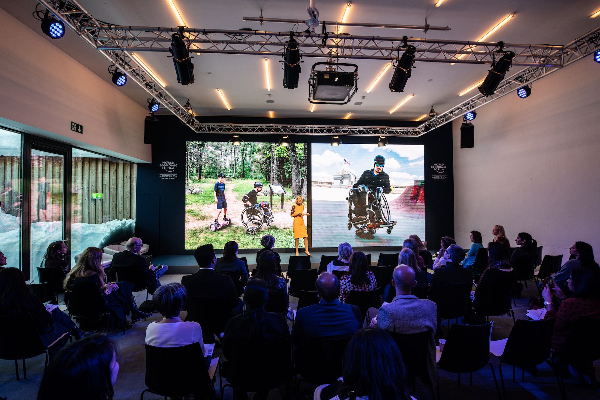 A woman in a mustard yellow dress stands in front of an audience, appearing to give a presentation. Behind her is a large screen showing people navigating in wheel chairs.