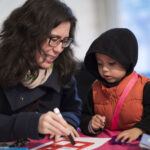 An adult and young child are sitting together at a table. The light skinned adult with long black hair and glasses is holding a marker and they demonstrate how to draw a square using a template. The child has light toned skin, wears a black long sleeve jacket with a hood over their head, and an orange puffer vest over the top.