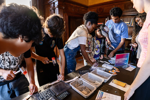 A photograph of a group of teenagers standing around a table making leather key chains. Two teenagers on the left are hammering their key chains. In front of the teenagers, on the table are baskets filled with various tools and materials.