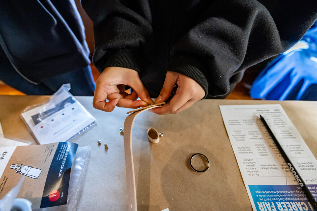 Two light brown hands pierce a metal stud through a leather strap. On the table sits an instruction manual, a key ring, metal studs, a worksheet, and a pencil.