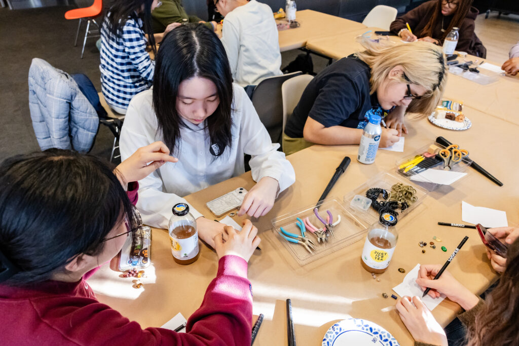 A photograph of a group of teenagers sitting at a table creating jewlery. The teenager on the left reaches their arm across and measures the finger of the teenager in front of them. At the center of the table are trays of pliers, wire, beads, pencils and scissors.