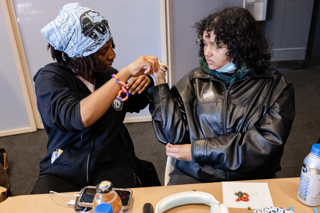 A photograph of a teenager with light brown skin, black curly long hair, and wearing a black leather jacket holds a spool of gold wire. A dark-skinned teenager on the left who has dreadlocks and wears a bandana pulls the wire.