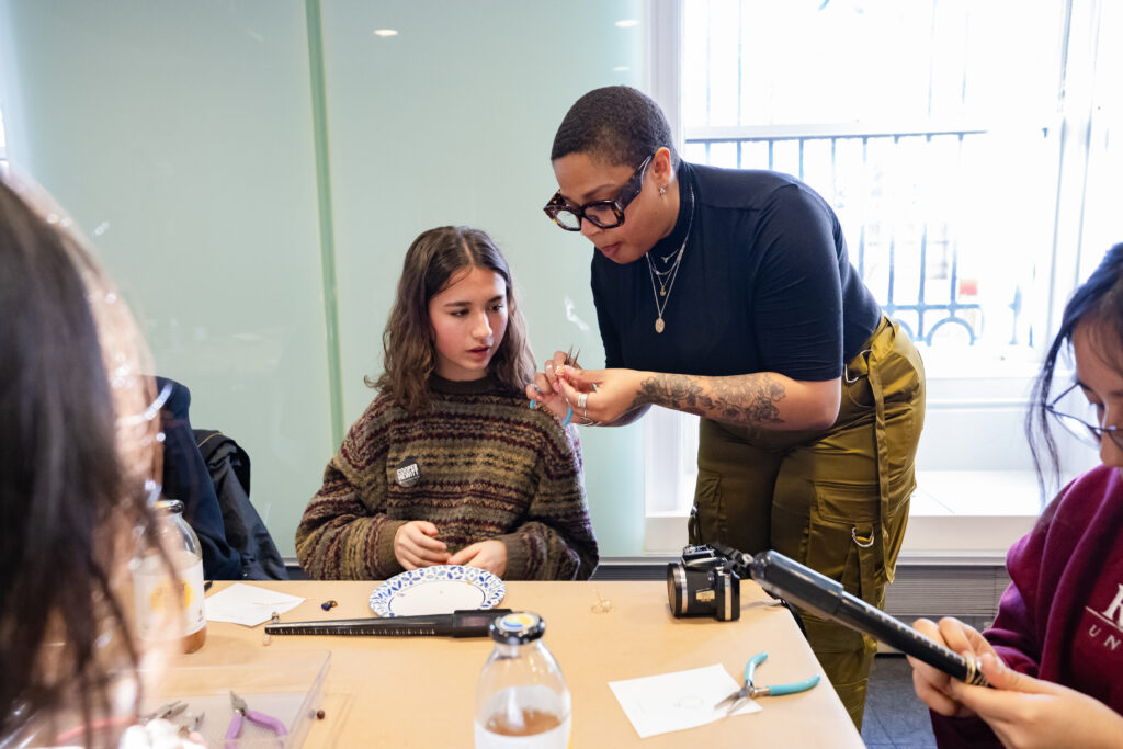 A photograph of an adult with light brown skin, closely shaved hair, and thick-framed glasses leans over to demonstrate an activity to a light-skinned teen with shoulder-length brown hair who is sitting at a table in front of them.