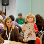 A photograph of a young light skinned child with curly blond hair holding up a piece of paper that has their t-shirt design, which has many different coloured scribbles all over. A light skinned, blonde haired adult sits beside the child smiling and helping the child hold up the drawing.