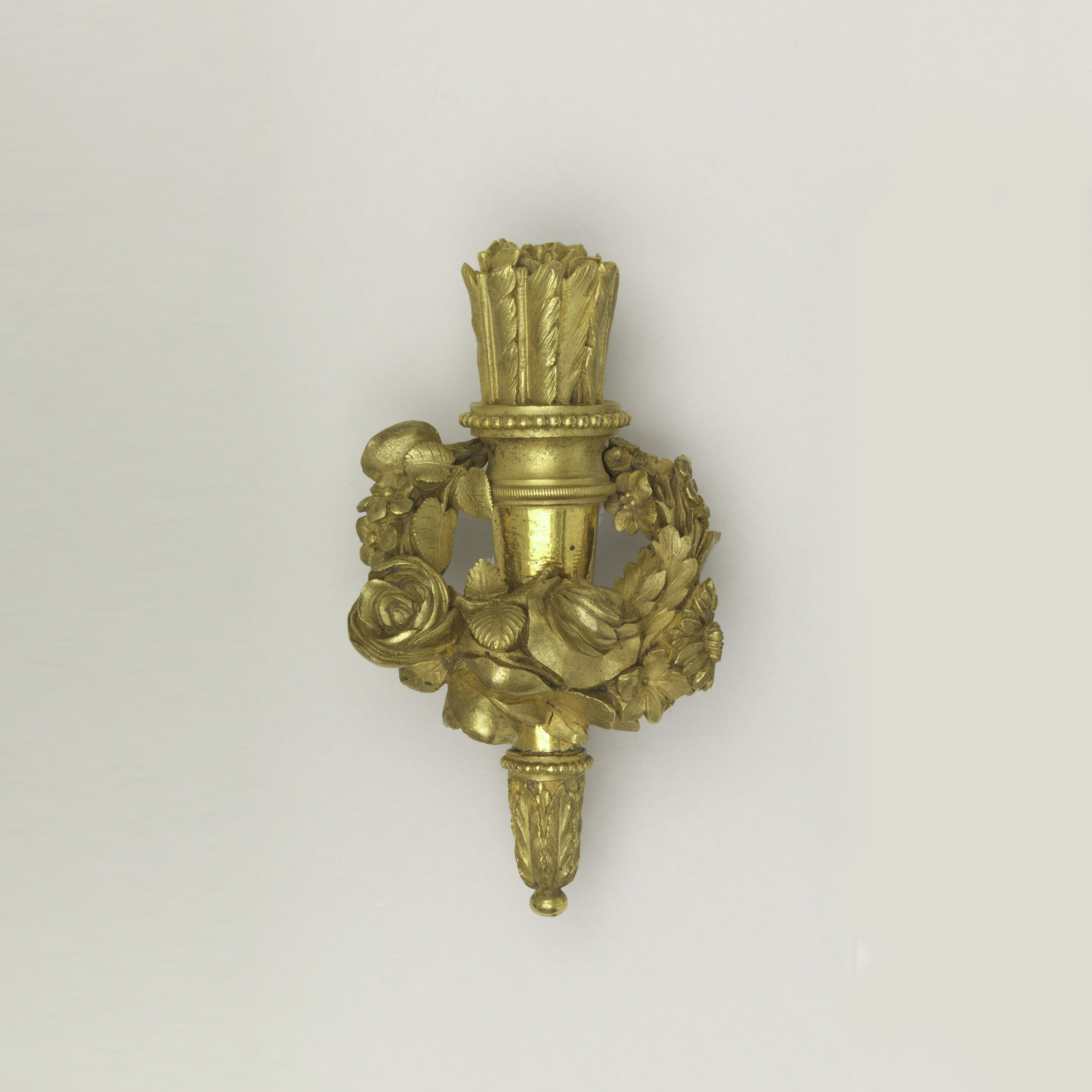 A gold, ornate sculptural object sits on a white surface, pictured from above. It features a quiver and arrows with a floral swag encircling it.