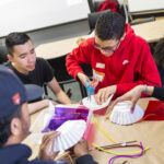 A photograph of a group of teenagers doing a craft activity. A medium skin toned teenager with short black hair and wears a red sweatshirt and glasses is cutting a paper coffee filter. Craft materials are scattered around the table.