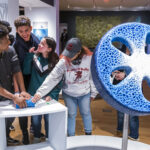 A photograph of a group of teenagers in a gallery space, gathered around a small blue object filled with fishnet-like holes. They smile playfully while reaching out to touch it. In front of the group is a large object on a stand that is around the shape and size of a car tire. It has the same blue colour and holes as the smaller object.