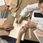 A photograph of a bird's eye view of two medium-skin toned teenagers sitting at a table and drawing on a notepad. They are drawing something in the shape of an envelope. Around them are materials such as paint brushes, leather straps and sheets, and worksheets on how to construct a leather accessory.