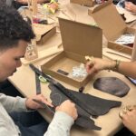 A photograph of two teenagers sitting at a table and making a leather accessory. Materials are scattered around the table such as hole punchers, leather pieces, scissors, threads, and cardboard boxes. The medium skin toned teenager sitting on the right with dark curly short hair is threading a flat piece of black leather.