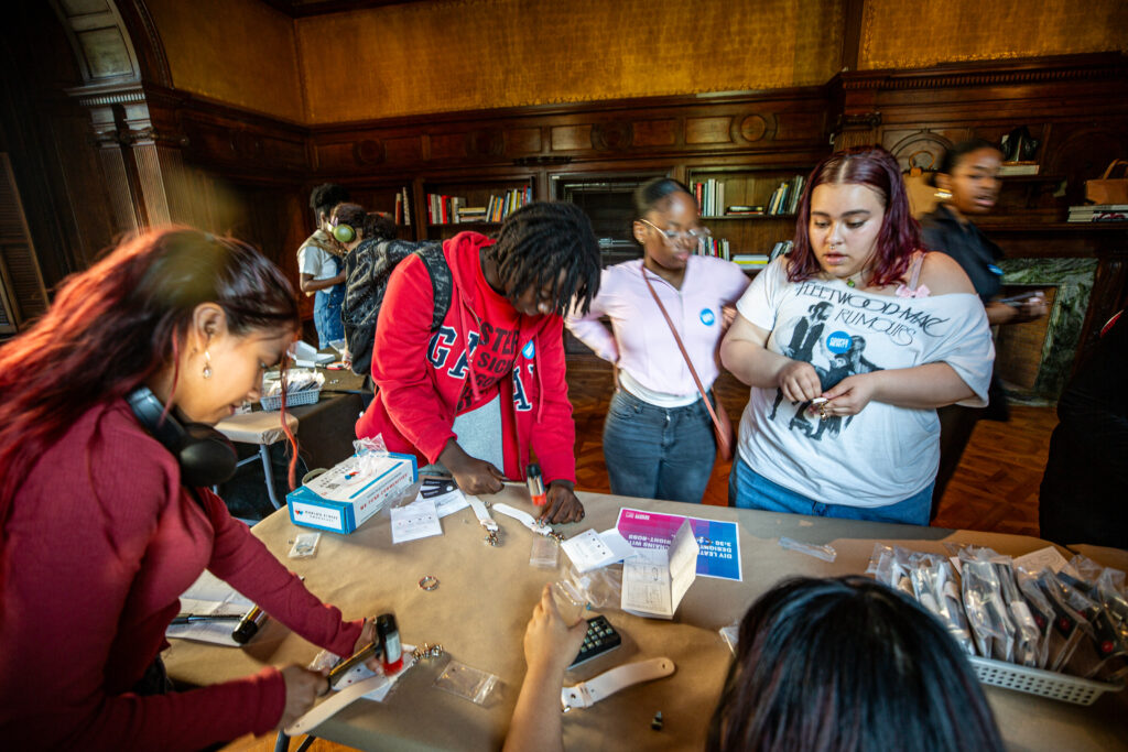 A photograph of a group of teenagers standing around a table making leather key chains. Two teenagers on the left are hammering their key chains. In the background there are shelves filled with books.