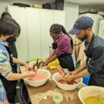 A photograph of a group of teenagers standing around a table each dipped a piece of clamped folded fabric into a bowl of liquid. Each teenager is wearing an apron and in front of each one of them is a bowl of colored liquid such as mustard, brown, and pink.