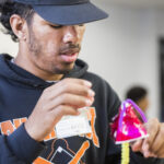 A photograph of a medium-dark skinned teenager wearing a black and orange baseball cap and sweatshirt who constructs something that looks like a desk lamp made out of paper cups, purple and yellow pipe cleaners, and pink paper.