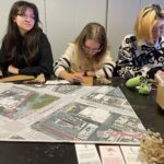 A photograph of a group of teenagers sitting at a table each drawing on a piece of triangular cardboard, a city map is laid out on the table in front of them. A section the same size and shape as their cardboard pieces is outlined in pink on the map. In the foreground of the photo are three white cards labelled local employee, bees, and pet owner. On the table there are other materials such as markers and dried twigs.