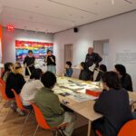 A photograph of a large group of teenagers sitting on bright orange chairs around a long table filled with printed out images and pages of text. At the far end of the table stands three adults, the adult on the left is smiling at the group of teenagers and the adult in the center is speaking to the teenagers. Behind the group in the right corner of the room there is a wall of multiple rows of brightly colored images, that are arranged in a color sequence starting from red to orange, yellow, blue, purple, pink, black, and white.