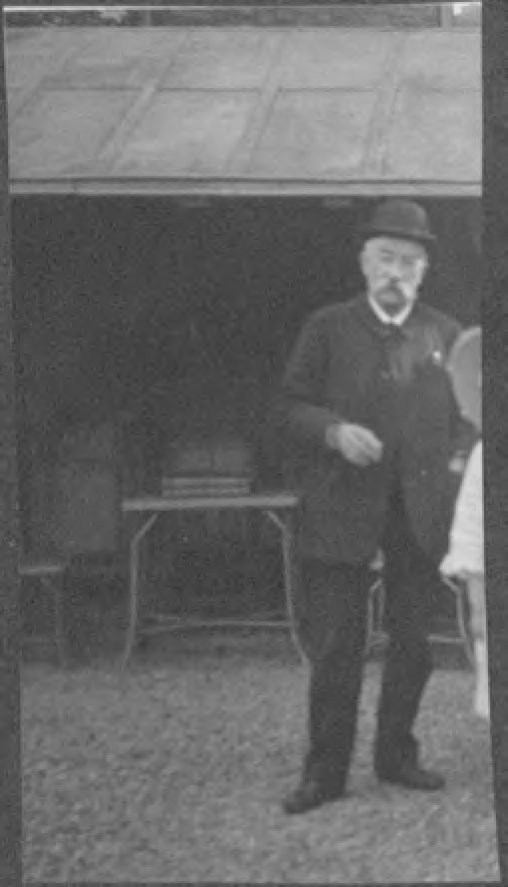 Black-and-white photograph depicting a person dressed in a dark suit, wearing a rounded hat. The person is standing toward the right of the image in front of a shed-like structure that is open; inside stands a table supporting a stack of books.