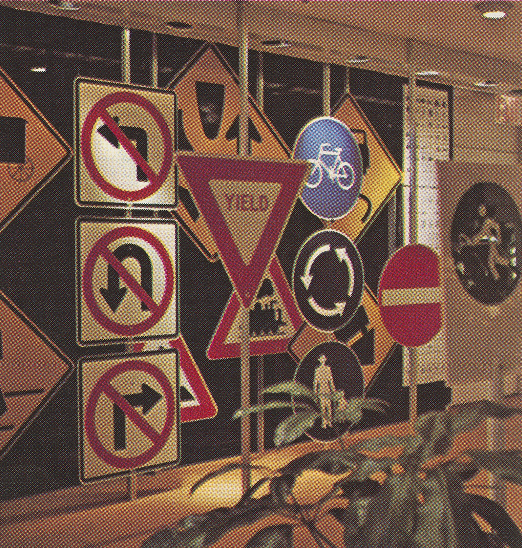 Photograph in color of an interior with a series of large street signs with symbols arranged in a display space. Lights illuminate them. In the foreground is a plant toward the bottom right corner.