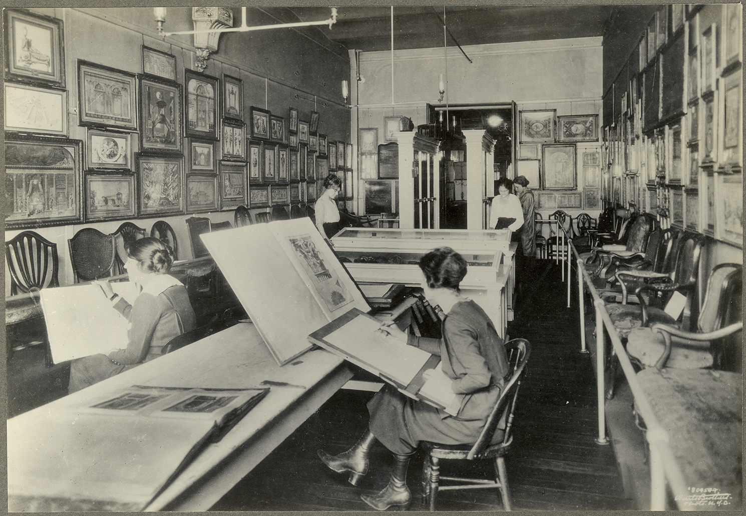 Black-and-white photograph of a large room filled with objects. Framed works hang side by side on the walls, entirely filling them. Different chairs sit along the entire length of the walls. A long table is in the center of the room with large books. Several people are engaging with the space either standing or sitting by the books.