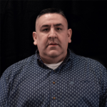 Portrait of Chief Brad KillsCrow who is wearing a blue and white patterned shirt in front of a black background.