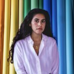 Portrait of Mona Chalabi who is wearing a white shirt and has long hair in front of a multicolored curtain.