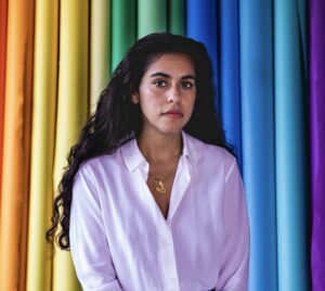 Portrait of Mona Chalabi who is wearing a white shirt and has long hair in front of a multicolored curtain.