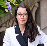 Portrait of Maria Nicanor who has long brown hair and is wearing red glasses, a white jacket and black shirt.