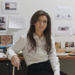 Color portrait of Amie Siegel. She has long, brown hair and sits while leaning against a desk. There are notes and sketches hanging on the wall behind her.