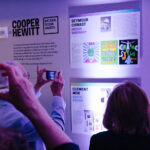 3 people observing and taking photos of information about the 2023 National Design Award Honorees posted on a wall. There are 4 profiles visible in the image: Seymour Chwast, BioCemenet Tiles by Biomason, Clement Mok, and Naeem Khan.