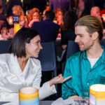 2 people in festive attire converse at a dining table. More dining tables and guests are in the background.