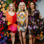 3 women dressed in festive, formal dresses pose in front of a bright and colorful floral backdrop.