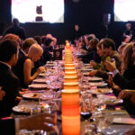 A long dining table in an event hall with people seated on each side. Dinnerware and lamps line the center of the table. Some guests applaud while others converse. A stage is visible in the background.