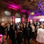 A crowd of people dressed in formal attire fills a large room with high ceilings. Pink and purple lights illuminate the detailed crown molding of the ceiling and 2 chandeliers.