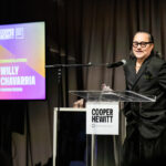 Willy Chavarria stands at a podium speaking into a microphone. He is in front of a black backdrop. A screen on the left side of the image shows his photo and text that reads: Cooper Hewitt National Design Awards, Congratulations Willy Chavarria, Fashion Design.