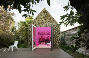 A group of people sit in casual conversation inside of a modern one-room, pink-lit structure with large glass doors that open onto a deck.