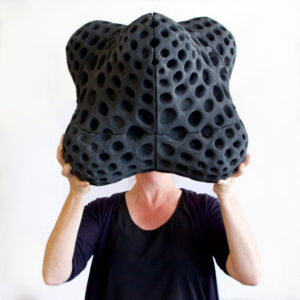 A person holds in front of their face a head-sized charcoal-colored object covered in exaggerated sponge-like holes.