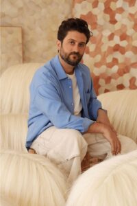 A person wearing an open blue button-down shirt over a white t-shirt, sitting amongst furry stools.