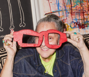 A person smiling and holding up a pair of whimsical, oversized, red eyeglasses extending beyond their face, with colorful and monochromatic artwork in the background.