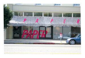 Photograph of a building facade with the word [PAPER] in hot pink capital letters splashed across its awning and windows.