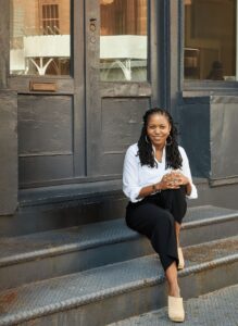 A person smiling, wearing a white top and black pants sitting on steps at the entrance of a building.