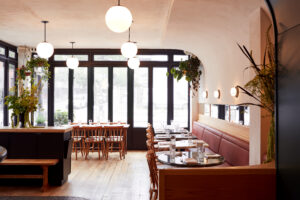 A midcentury modern restaurant interior with orb-shaped lighting, large street-facing windows, blush leather booth and wooden table seating, and decorative floral arrangements.