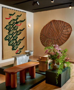 A design fair booth interior with wall mounted tapestries and a display of three carved benches and a floral arrangement on dark green pedestals.