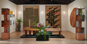 A design fair booth interior framed by modular bookshelves with floral arrangements, large geometric wall hangings, framed prints, and a trio of carved benches.