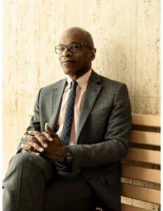 A person wearing glasses and a suit and tie, sitting on a bench.