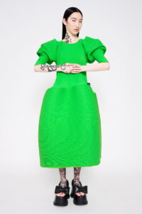 A heavily tattooed model wears a boxy, bright green short-sleeved dress and black platform shoes with large bows.