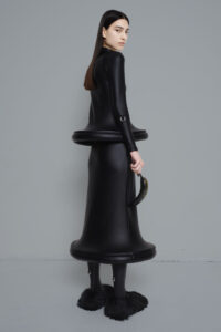 A model wears a form-fitting shiny black dress in the shape of two bells stacked on top of one another.