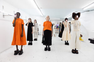 A group of models ranging in age and skin color stand in three perfect rows wearing haute couture garments of white, black, and orange and hold smartphones up to their faces displaying another face than their own.