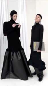 Two models are casually posed together wearing long black drop-waist garments.