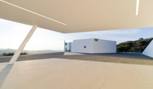 Exterior view of an angular pavilion overlooking a steep bluff with a white heptagonal building in the distance on the corner edge of the cliffside.