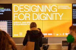 People in front of a large screen from the deem symposium that reads [Designing for Dignity] in towering capital letters.